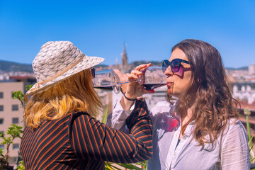 A lesbian couple with their arms linked and holding glasses of red wine, drinking them together on a rooftop in Barcelona overlooking the Sagrada Familia on a sunny day with a clear blue sky.