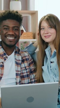Smiling young two multiethnic man and woman sitting on couch at home during previsioning interesting program on laptop and posing on camera.