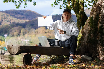 Casual observer holding and indicating blank sheet on outdoor wooden seat amid fall foliage and...