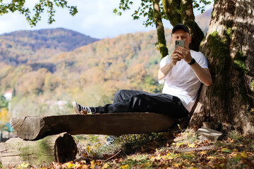 Fototapeta premium Seated male using phone camera for selfie in vibrant woodland with mountain horizon