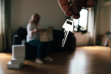 Hand holding apartment keys with person unpacking moving boxes in background.