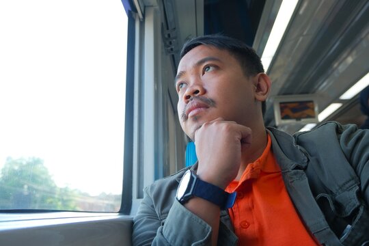 A Southeast Asian man sits by a bright train window, resting his chin on his hand while gazing outside, creating a reflective and peaceful travel moment.