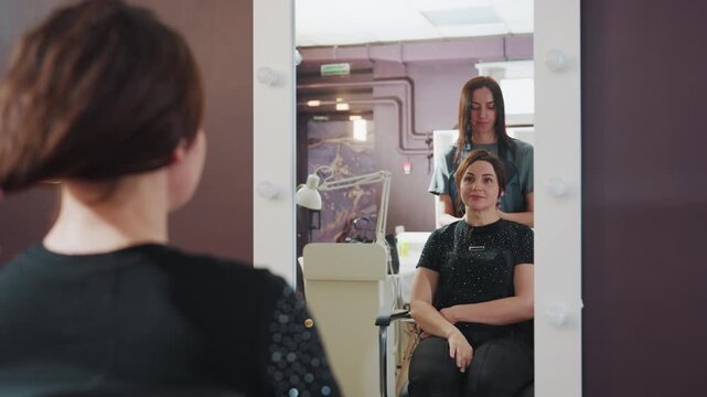 caucasian woman facing mirror in salon tying ponytail while stylist observes, calm consultation mood, visible product shelves and contemporary interior, client deciding haircut length