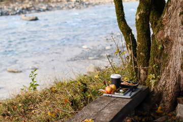 Apple, coffee cup, snacks on notepad by river under tree