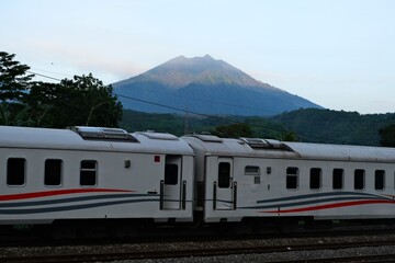 White passenger train with red and gray stripes travels along tracks with a scenic backdrop of a forested mountain under clear skies.
