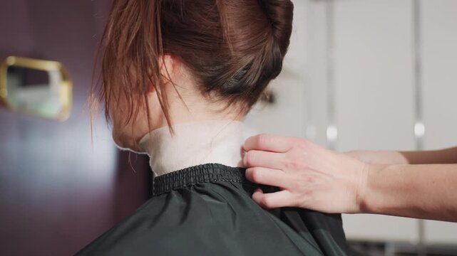 secure protective barrier, stylist attaches cape and neck strap, client reclines with hair lifted in calm environment, relaxed client with stylist securing cape under salon lighting conditions