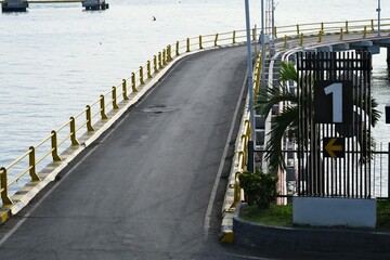 Paved road with yellow railings curves toward a ferry terminal gate over calm coastal waters, surrounded by signs, lights, and distant hills.
