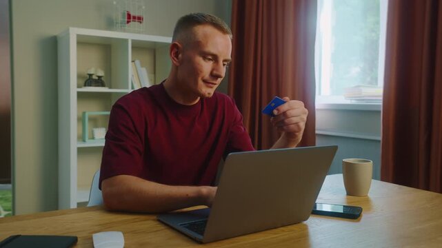 Young adult man sit desk home pay by bank credit card buy stuff online use laptop computer, mobile cell phone. Smiling person do purchase internet store. Remote shopping. Big holiday sale, discounts.