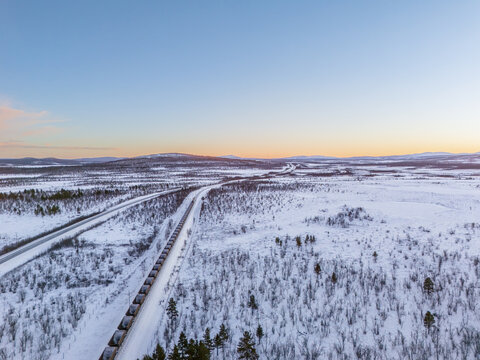 Aerial view of a long, dark iron ore train snaking through the snowy landscape, contrasted against the soft hues of the horizon in winter, Kiruna, Norrbotten County, Sweden.