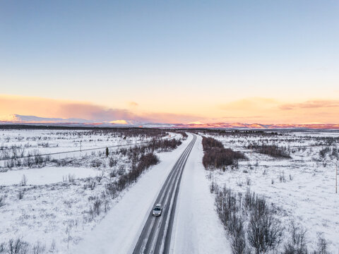 Aerial view of a lone car driving down a snow-covered highway, framed by a pastel sky and distant mountains, Kiruna, Norrbotten County, Sweden.