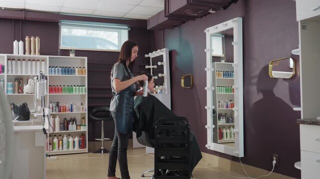 caucasian stylist arranging salon chair and tools, setting up mixing station and product shelves, wide interior shot showing tidy equipment, organized trolley and mirror, professional pre-appointment