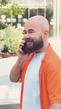 Portrait of good-looking satisfied successful young man with beard which going down on wide street with bike and has phone conversation.