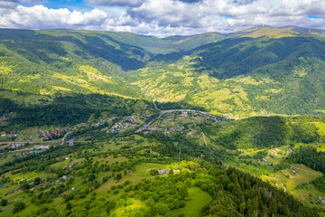 A high-altitude aerial view of a scenic mountain valley with lush green hills, forests, and scattered rural homes, showcasing a peaceful natural landscape under a dramatic cloudy sky.