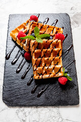 Sweet and crispy waffles sit on a dark slate, accented by fresh berries and chocolate sauce