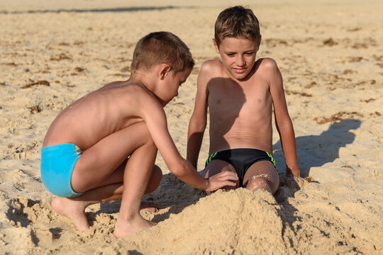 Two boys are playing in the sand. Brother buries brother with sand on the beach