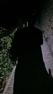 Shadow, person, walking up a dark mysterious narrow path next to a stone wall at night Vertical video