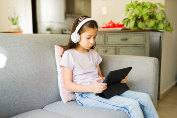 Little girl wearing wireless headphones and using a digital tablet at home, engaging in e-learning and online education