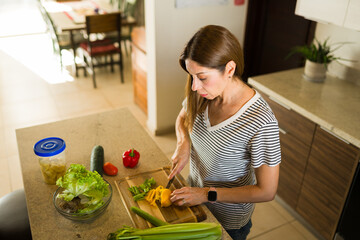 Adult woman chopping yellow bell pepper on a wooden board, preparing fresh vegetables for a healthy salad in kitchen