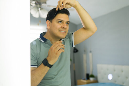Smiling man while combing his hair in front of a mirror, performing a daily grooming routine in a bedroom