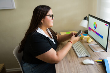 Plus size woman working from home as graphic designer, creating a color palette using smartphone and desktop computer