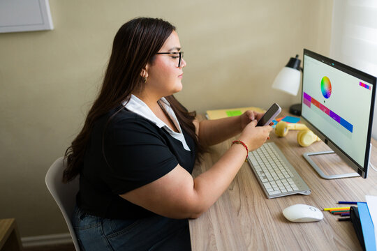 Plus size woman working from home as graphic designer, creating a color palette using smartphone and desktop computer