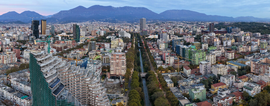 A wide aerial panorama of Tirana featuring the Lana River, dense city neighbourhoods and mountains in the distance. - Powered by Adobe