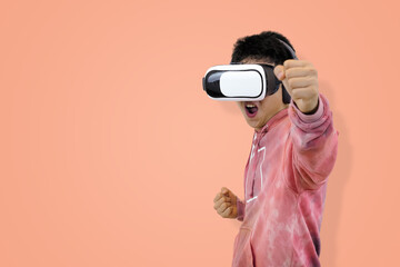 Triumphant young Asian man in hoodie wearing a virtual reality headset, cheering with a raised fist, symbolizing victory or success in gaming in studio background