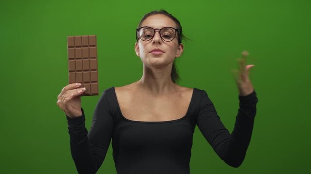 Woman holds large chocolate bar in left hand and flips hair with right hand in green studio set; playfulness indulgence confidence.