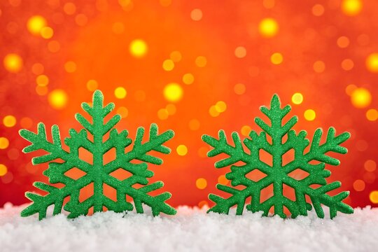Two green snowflake decorations sitting in snow against a blurry orange background