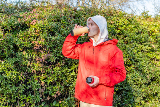young male runner in sportswear taking break during morning run drinking warm beverage, hot drink from wooden thermos, self care and wellness, feeling energized, outdoors healthy and active lifestyle