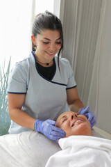 Obraz premium A young esthetician in uniform performing a professional facial treatment on a mature female client in a clean spa environment.