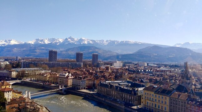A stunning, wide view of Grenoble's urban sprawl set against the dramatic, snow-capped peaks of the Belledonne mountain range. The Is&egrave;re River and the Three Towers stand out.