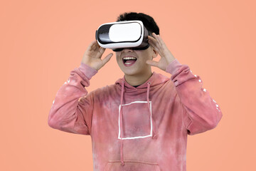 Excited young Asian man wear hoodie reacting with joy while wearing a virtual reality headset, symbolizing the thrilling first experience in studio background