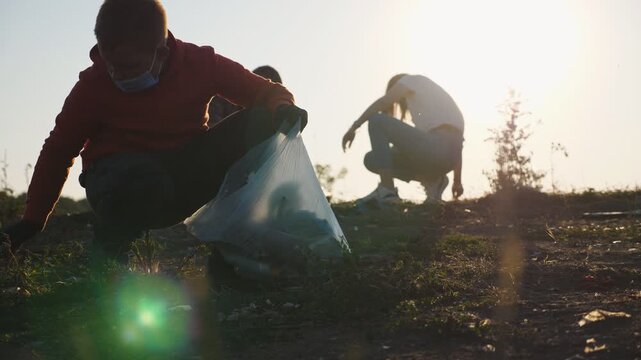 Close up of little male kid in gloves collect trash in bag saving nature at countryside. Parents of boy help to cleaning lawn of paper waste at background. Concept of environmental problem. Slow mo