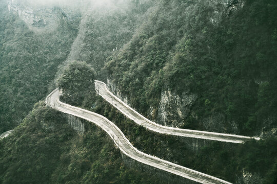 View of a winding mountain road cuts through verdant hills under a misty sky, showcasing nature's grandeur and human engineering's ambition, Zhangjiajie, China.
