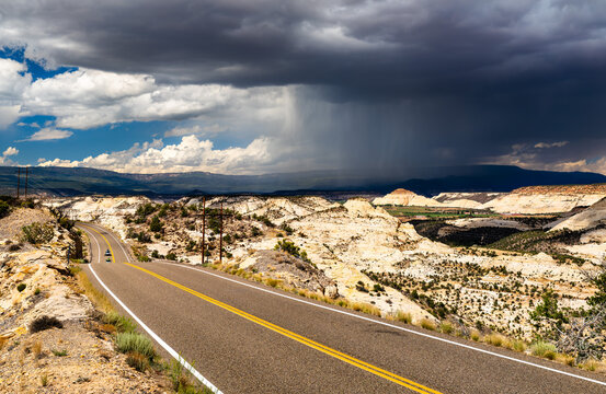 Utah State Route 12 winds over The Hogback in Grand Staircase-Escalante National Monument, United States. A large, dramatic storm cloud with rain, virga, hangs over the vast slickrock landscape
