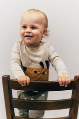 Portrait of a one-year-old child with beautiful curly hair standing on a chair by a white wall and smiling brightly, creating a warm and joyful atmosphere.
