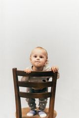 Portrait of a one-year-old child with beautiful curly hair standing on a chair by a white wall and smiling brightly, creating a warm and joyful atmosphere.