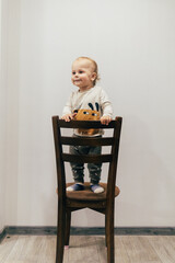 Portrait of a one-year-old child with beautiful curly hair standing on a chair by a white wall and smiling brightly, creating a warm and joyful atmosphere.