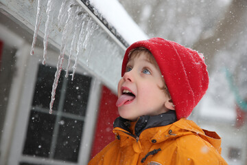 Boy child in red hat and jacket tasting an icicle in falling snow, winter outdoors scene with curious gaze and playful tongue moment of childhood wonder in cold weather
