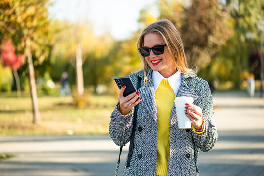 Portrait of urban businesswoman with sunglasses in stylish coat using smartphone and drinking coffee while standing in the city park during sunny autumn day. - Powered by Adobe