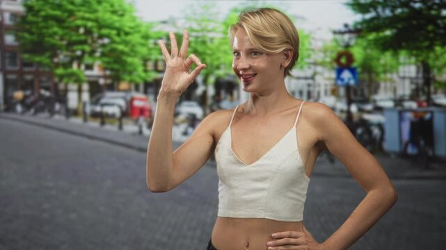 Caucasian blond woman shows ok with hand and puckered lips on street in urban cobblestone scene wearing cami and casual top; playful confidence.