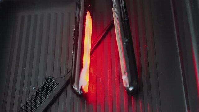 close view of warm styling device preparing, brightly illuminated hair straightener heating up on platform, closeup shot of glowing red straightener and comb awaiting use in salon environment