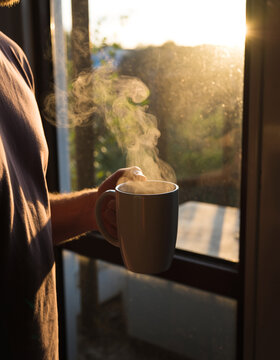 una persona sostiene un caf&eacute; cremoso y humeante mientras observa un bonito atardecer a trav&eacute;s de una ventana. La luz c&aacute;lida del sol crea un ambiente tranquilo y acogedor.