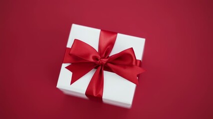 A white gift box tied with a vibrant red satin ribbon bow rests on a red surface - Powered by Adobe