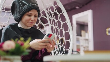 woman seated under hair dryer using phone in salon; quiet waiting moment with cape on, muted colors and floral foreground, scrolling through messages while stylist attends other client
