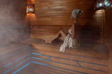 Young woman relaxing and sweating in hot sauna wrapped in towel. Girl In Sauna. Interior of Finnish sauna, classic wooden steam room with hot steam. Russian bathroom. Relax in hot Bathhouse with steam