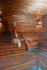 Young woman relaxing and sweating in hot sauna wrapped in towel. Girl In Sauna. Interior of Finnish sauna, classic wooden steam room with hot steam. Russian bathroom. Relax in hot Bathhouse with steam
