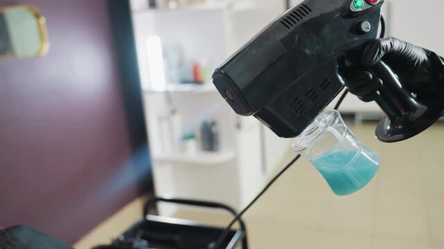 handheld misting device emitting fine vapor across salon chair, blue solution bottle attached, technician operating trigger with gloved hand, modern salon interior and gentle treatment atmosphere