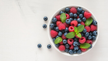 Bowl of fresh blueberries and raspberries with mint leaves on white wooden background. Healthy mix of berries for breakfast or snack.